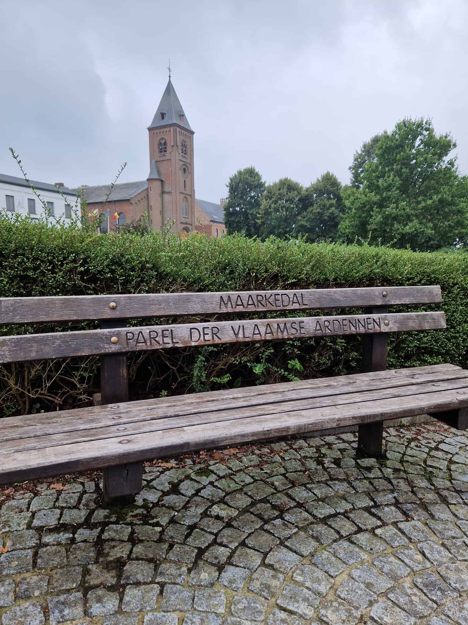 Houten zitbank met het opschrift Maarkedal, parel der Vlaamse Ardennen, met de kerk van Louise-Marie op de achtergrond